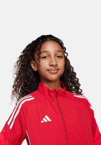 Rode zip-up jas met witte strepen op de schouders, gemaakt van een stof met een lichte textuur, voorzien van een Adidas-logo op de borst.