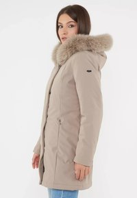 Piumino beige con cappuccio in pelliccia sintetica, due zip laterali, maniche lunghe e taglio dritto. Tessuto liscio e materiale resistente all'acqua.
