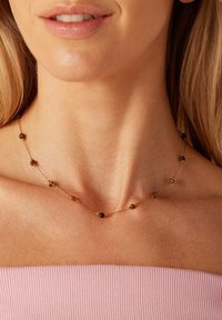 Gouden ketting met ronde kralen in bruin- en zwarttinten, gelijkmatig verdeeld over de lengte, tegen een lichtroze top.