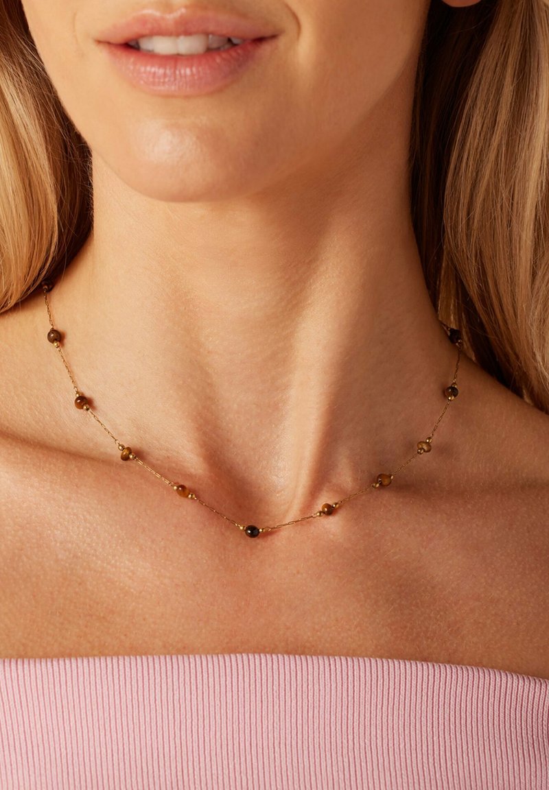 Gouden ketting met ronde kralen in bruin- en zwarttinten, gelijkmatig verdeeld over de lengte, tegen een lichtroze top.