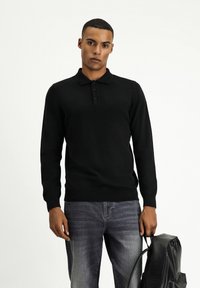 Schwarzes Langarmpolo mit geripptem Kragen und Knopfleiste mit drei Knöpfen, kombiniert mit ausgewaschenen Jeans und einem schwarzen Leder-Rucksack.