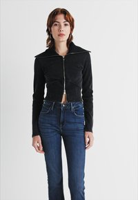 Chaqueta negra corta con cierre frontal, cuello puntiagudo y tela texturizada; combinada con jeans de tiro alto en azul oscuro.