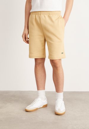Pantalones cortos de algodón beige con una cinturilla elástica, bolsillos laterales y un parche de logo verde, combinados con zapatillas blancas y calcetines cortos.