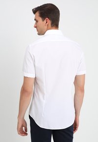 Camicia bianca a maniche corte con colletto dritto, dettaglio di spalla sul retro e orlo curvato, realizzata in tessuto morbido senza motivi visibili.