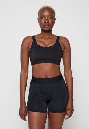 Femme aux cheveux blonds courts portant un soutien-gorge de sport noir et un short de sport noir ajusté, debout devant un fond clair uni.