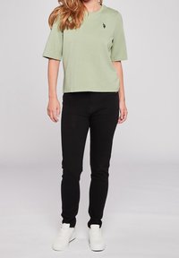 Žena v světle zeleném tričku s krátkým rukávem, užších černých kalhotách a bílých teniskách stojící na jednobarevném světlém pozadí.