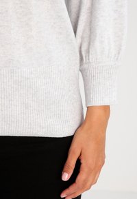 Hellgrauer Pullover mit gerippten Bündchen und Saum, aus weichem, strukturiertem Stoff. Zeigt eine Hand vor einem schwarzen Hintergrund, der die Detailgestaltung des Ärmels hervorhebt.