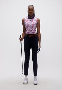 Γυναίκα με αμάνικη πόλο μπλούζα, σκούρο παντελόνι και γάντι γκολφ κρατάει ένα μπαστούνι γκολφ, στέκεται μπροστά σε απλό λευκό φόντο.