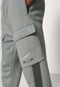 Grå sweatpants har en sidoficka, silvrig Nike-logga och en kontrasterande svart rand längs benet. Mjuk textur och avslappnad design.