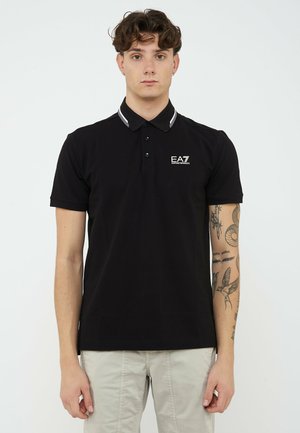 Giovane uomo con polo nera EA7 Emporio Armani con rifiniture bianche e pantaloni chiari, in piedi con espressione neutra e tatuaggi visibili sulle braccia.