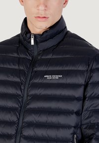 Giacca a vento blu con design trapuntato, colletto alto e zip. Presenta il logo di Armani Exchange sul petto. Texture liscia e lucida.