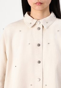 Camicia in denim beige chiaro con colletto a punta, bottoni a pressione argentati e borchie metalliche posizionate uniformemente sul tessuto.
