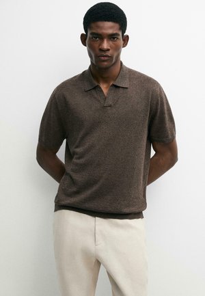 Mężczyzna stoi z rękami za plecami, ubrany w brązowe krótkie dzianinowe polo i jasnobeżowe spodnie na jednolitym białym tle.