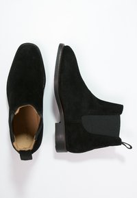 Bottes de cheville en daim noir avec une finition lisse, des côtés élastiques et un talon empilé bas. Comprend une languette à l'arrière pour un enfilage facile.