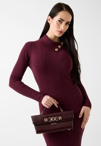 Vestito a costine color marrone scuro con collo alto decorato da accenti dorati. La modella tiene in mano una borsa marrone scuro con accessori dorati e il nome del marchio visibile.