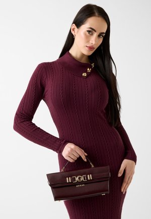 Vestito a costine color marrone scuro con collo alto decorato da accenti dorati. La modella tiene in mano una borsa marrone scuro con accessori dorati e il nome del marchio visibile.