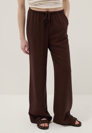 Persoon die losse donkerbruine broek met trekkoord en donkerbruine sandalen met metalen studs draagt, staande tegen een effen achtergrond.