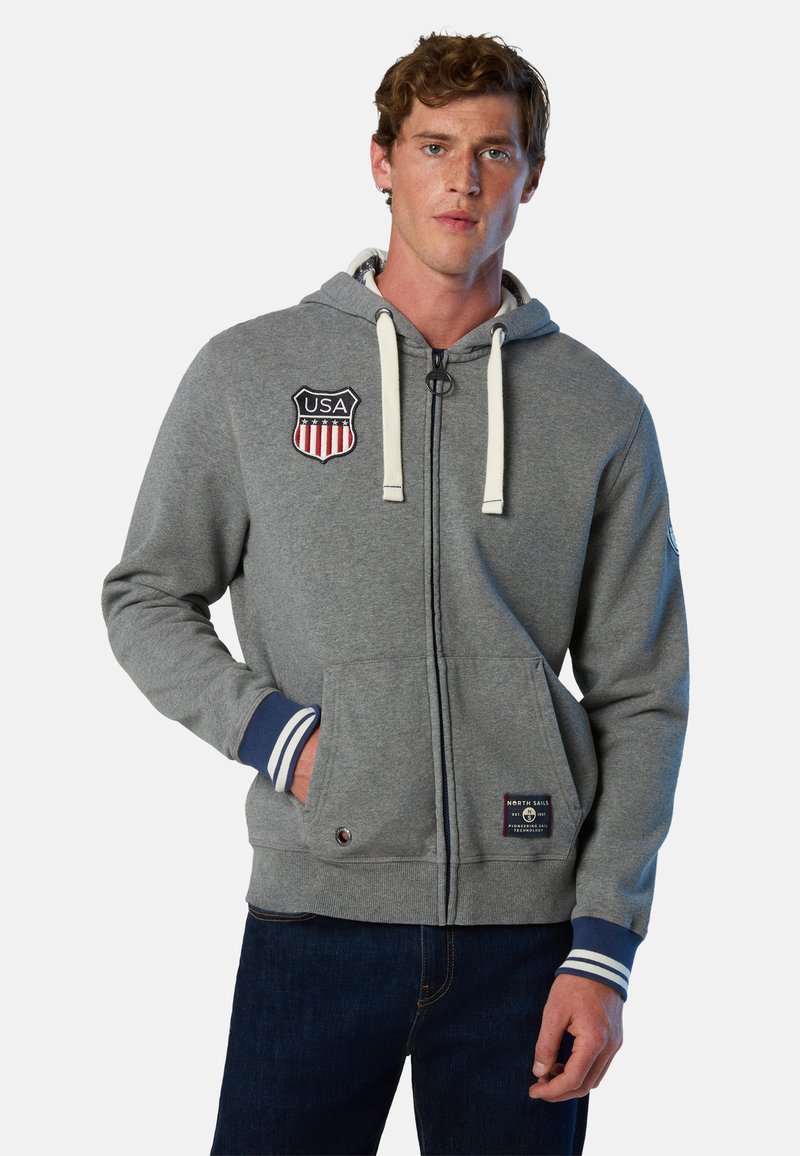 Felpa grigia con zip e toppe degli Stati Uniti, con cordini, tasche frontali e polsini a righe. Realizzata in materiale morbido con una vestibilità casual.