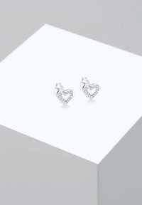 Heart-shaped silver earrings featuring a hollow center, accentuated by a rim of clear stones, resting on a minimalist display.