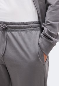 Κοντινό πλάνο ατόμου που φορά γκρι φόρμα με ελαστική μέση και κορδόνι, χέρι στην τσέπη, σε συνδυασμό με γκρι μπουφάν και λευκό πουκάμισο.