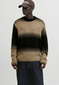 Eine Person trägt einen gestrickten Pullover in Braun- und Schwarztönen, dunkle weit geschnittene Jeans, eine braune Mütze und kleine Creolen und steht vor einem schlichten Hintergrund.
