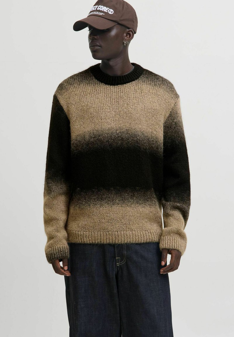 Eine Person trägt einen gestrickten Pullover in Braun- und Schwarztönen, dunkle weit geschnittene Jeans, eine braune Mütze und kleine Creolen und steht vor einem schlichten Hintergrund.