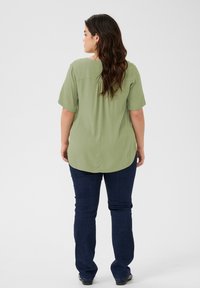 Top verde chiaro a maniche corte con vestibilità rilassata, pieghe posteriori e orlo arrotondato, abbinato a jeans blu scuro a gamba dritta. Texture liscia.