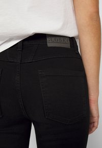 Jeans de denim negro con un diseño ajustado, que presentan un parche de logo de plástico en la cintura y dos bolsillos traseros con detalles de costura.
