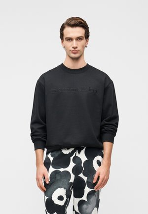Μαύρο φούτερ με ανάγλυφο κείμενο "marimekko", στρογγυλή λαιμόκοψη και μακριά μανίκια· συνδυασμένο με μαύρο-λευκό μοτίβο παντελόνι με floral σχέδιο.