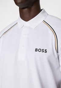 Chemise polo blanche à col, avec un logo noir et de fines rayures noires et dorées le long des épaules. Détails en maille sur le col.