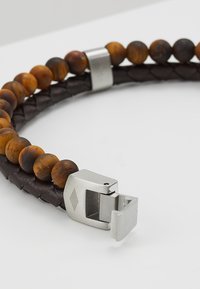 Bracciale in pelle con texture intrecciata marrone, con perline di legno alternate in tonalità di ambra e una chiusura in metallo argentato con un accento di diamante.