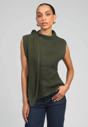 Femme à la peau foncée portant un haut tricoté vert olive sans manches avec un détail d'écharpe attachée, un jean foncé, et des boucles d'oreilles créoles en or, posant avec une main sur la hanche.