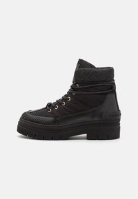 Bota negra de caña alta con suela gruesa, cuello acolchado y de textura acolchada, cordones redondos pasados por ojales dorados y puntera texturizada sobre fondo blanco.