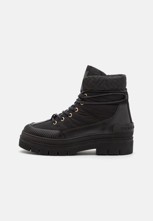 Botte montante noire avec semelle épaisse, col rembourré matelassé, lacets ronds passés à travers des œillets dorés, et capuchon de nez texturé sur fond blanc.