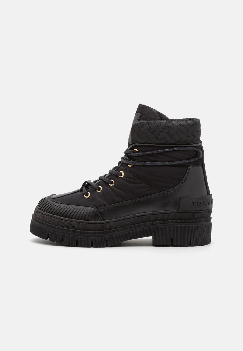 Bota negra de caña alta con suela gruesa, cuello acolchado y de textura acolchada, cordones redondos pasados por ojales dorados y puntera texturizada sobre fondo blanco.