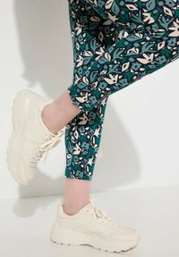 Scarpe da ginnastica bianche con suole testurizzate e lacci, abbinate a leggings attillati color teal con un motivo floreale in crema e verde scuro.