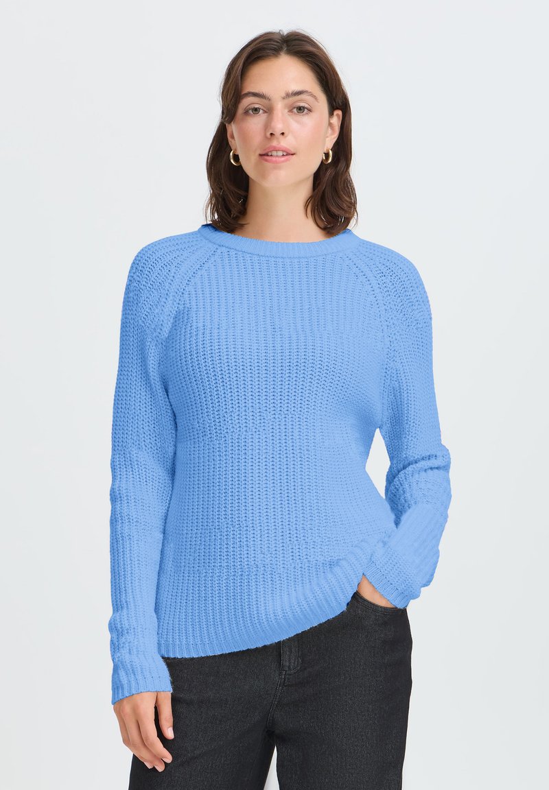 Femme aux cheveux foncés portant des boucles d'oreilles en or en forme d’anneaux, un pull tricoté texturé bleu clair et un pantalon noir, debout avec une main dans la poche.