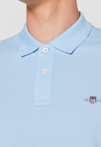 Polo bleu clair avec deux boutons blancs fermés, un col et un petit logo brodé en forme de bouclier rouge, blanc et bleu sur la poitrine.