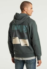 Felpa con cappuccio verde scuro con un motivo grafico sul retro che presenta tre blocchi colorati e testo. Materiale in cotone misto, morbido al tatto e dalla vestibilità ampia.