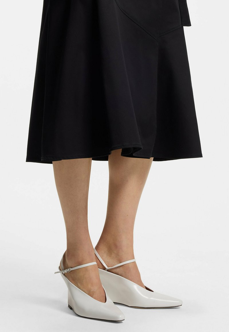 Jambes portant des chaussures compensées blanches à bout pointu avec de fines brides à la cheville, associées à une jupe noire évasée mi-longue.