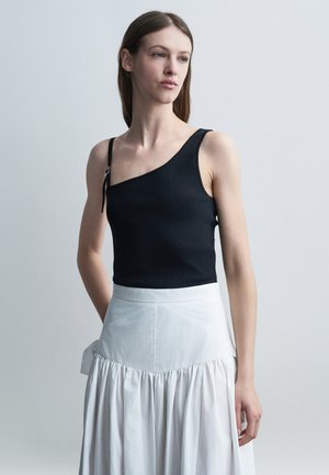 Kvinde i en sort enkelt skulder-top med stropdetalje og hvid høj-taljet nederdel, stående mod en ensfarvet lys baggrund.