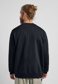 Felpa nera a girocollo, realizzata in cotone, presenta una vestibilità rilassata, polsini e orlo a costine, e un'etichetta sul lato sinistro inferiore.