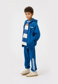 Felpa blu e pantaloni jogger realizzati in tessuto morbido, con strisce bianche e logo. Indossati con una maglietta a righe e sneakers beige.