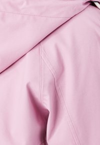 Jasnoróżowa kurtka z gładką strukturą, skośnym szwem na ramieniu i kapturem. Zauważalne detale przeszycia wzdłuż krawędzi.