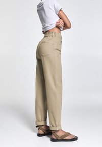 Pantalones de pierna ancha beige con cintura ajustable y puños enrollados, combinados con sandalias marrones de tiras, contra un fondo liso.