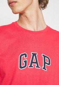 Persona vestida con una camiseta roja brillante con grandes letras azul marino y blancas "GAP" en el pecho, con la cabeza girada ligeramente hacia un lado.