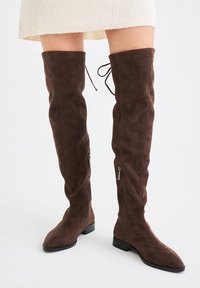 Botas altas de ante marrón con cremalleras laterales y cordones en la parte trasera, llevadas con una falda color crema sobre un fondo blanco.