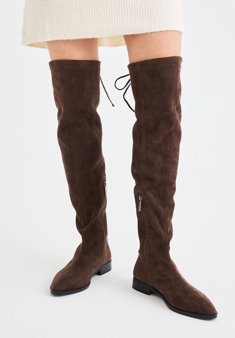 Botas altas de ante marrón con cremalleras laterales y cordones en la parte trasera, llevadas con una falda color crema sobre un fondo blanco.