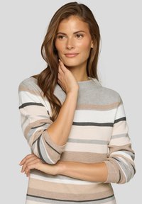 Frau mit langen braunen Haaren, die einen beigefarbenen, weißen und grauen gestreiften Pullover trägt, posiert mit einer Hand, die ihren Hals berührt, und dem anderen Arm verschränkt.