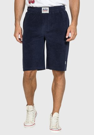 Uomo che indossa pantaloncini in spugna blu navy con vita elastica e sneakers bianche alte su sfondo chiaro.